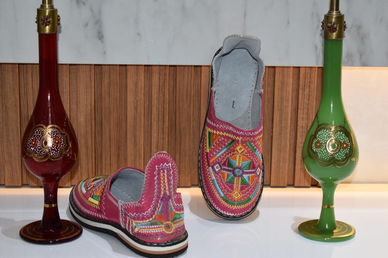 Puede incluir: Dos pares de zapatos rosas bordados sin cordones con patrones geom&eacute;tricos coloridos. Una jarra decorativa de vidrio rojo y verde con detalles dorados a cada lado de los zapatos.