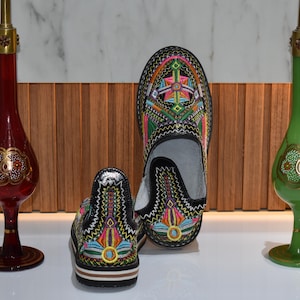 Puede incluir: Zapatos negros bordados con coloridos dise&ntilde;os geom&eacute;tricos. La imagen tambi&eacute;n presenta dos jarrones decorativos de vidrio, uno rojo y otro verde, cada uno con detalles dorados y una forma esbelta.