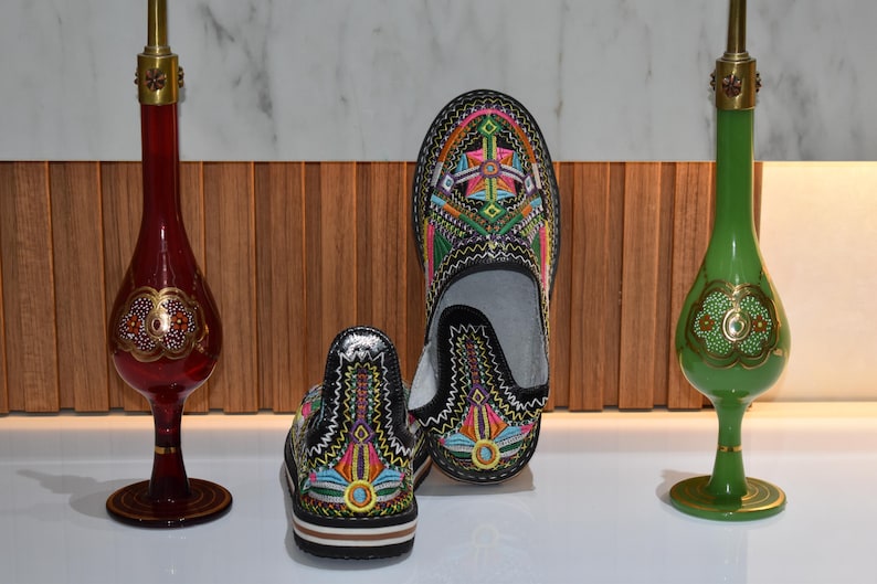 Puede incluir: Zapatos negros bordados con coloridos dise&ntilde;os geom&eacute;tricos. La imagen tambi&eacute;n presenta dos jarrones decorativos de vidrio, uno rojo y otro verde, cada uno con detalles dorados y una forma esbelta.