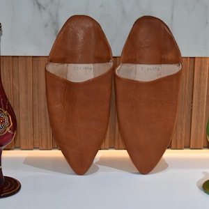 Könnte beinhalten: Zwei braune Lederschuhe mit spitzen Zehen werden zwischen zwei dekorativen Glasvasen präsentiert. Die Vasen sind rot und grün, mit goldenen und floralen Designs. Die Schuhe haben einen warmen, erdigen Farbton.