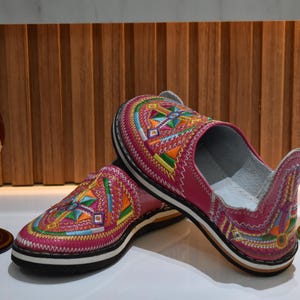 Puede incluir: Un par de zapatillas de cuero rosa con coloridos patrones geom&eacute;tricos bordados. Las zapatillas tienen una suela negra con una franja blanca. Jarrones decorativos de cristal en rojo y verde en el fondo.