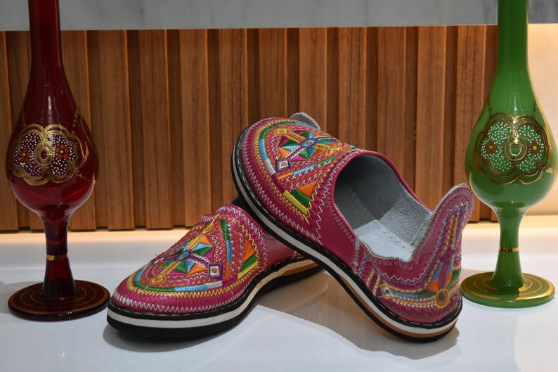 Puede incluir: Un par de zapatillas de cuero rosa con coloridos patrones geom&eacute;tricos bordados. Las zapatillas tienen una suela negra con una franja blanca. Jarrones decorativos de cristal en rojo y verde en el fondo.