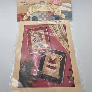 May include: A clear plastic packet containing the I Believe in Hollie quilting pattern by Cute as a Button presents Button Babies by Cheri, number 6251. The packaging features illustrations of two fabric wall hangings with holiday themes.