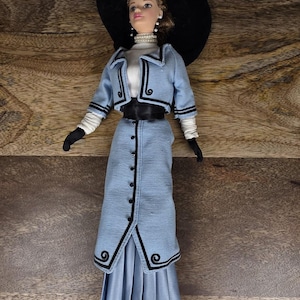 Puede incluir: Una muñeca vestida con una chaqueta azul claro y falda a juego con ribetes negros, una blusa blanca de cuello alto, guantes negros y un sombrero negro de ala ancha con una pluma blanca. La muñeca lleva un collar de perlas y pendientes.