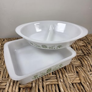 May include: Two white milk glass baking dishes featuring a green floral pattern. One dish is a square baker, and the other is a round divided serving dish with a central divider. Both pieces have smooth, glossy surfaces and integrated handles.