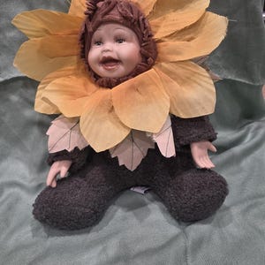 Puede incluir: Una muñeca caprichosa vestida de girasol. La muñeca tiene una cara de porcelana con mejillas rosadas y una expresión sonriente. Lleva una gorra marrón y un gran cuello de pétalos amarillos y naranjas. El cuerpo de la muñeca es marrón oscuro con detalles de hojas.