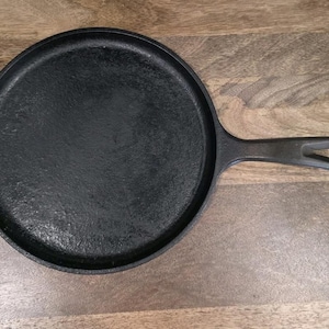 May include: A black cast iron griddle pan with a single handle. The pan is round with a flat cooking surface and a raised edge. The handle has a hole for hanging.