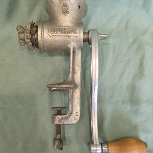 May include: A vintage Universal meat grinder made of silver-colored metal. The grinder has a hand crank with a wooden handle and a clamp for attaching to a table. The word "UNIVERSAL" is embossed on the side, and the number "2" is visible.