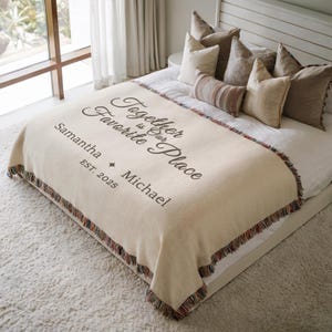 May include: A cream-coloured throw blanket with the text "Together is Our Favorite Place" and names "Samantha" and "Michael" with "EST. 2025". The blanket has a fringed edge with multiple colours. The blanket is on a bed with pillows.