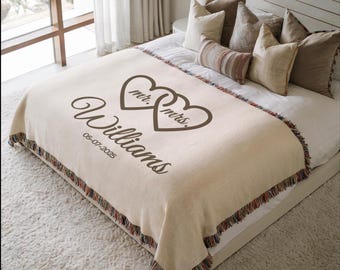 May include: A cream-colored woven throw blanket with fringed edges, featuring a dark brown graphic of two interlocking hearts containing the text mr. and mrs., the name Williams in script, and the date 05-07-2025 printed on the fabric surface.