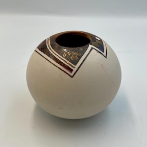 May include: A spherical, off-white ceramic vase with a small opening. The rim and upper portion of the vase feature a decorative band with geometric patterns in brown, gold, and red. The vase has a smooth, matte finish.