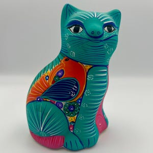 May include: A teal ceramic cat figurine with a hand-painted design. The cat has a smiling face, black eyes, and a colorful pattern on its body, including red, orange, and pink accents. The figurine is approximately 7 inches tall.