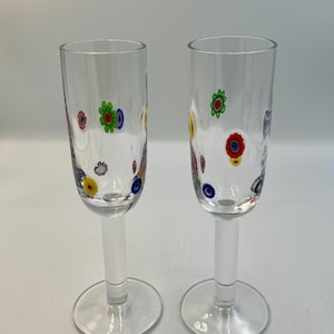 May include: Two clear champagne flutes with colorful floral designs. The flowers are in shades of red, yellow, blue, and green. The flutes have a tall, slender bowl and a clear stem and base. The glasses are perfect for a special occasion.