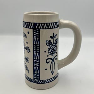 May include: A beige ceramic beer stein with a large handle. The stein is decorated with blue floral and geometric patterns. The top and bottom rims have a blue zigzag design. The floral designs include roses and leaves.