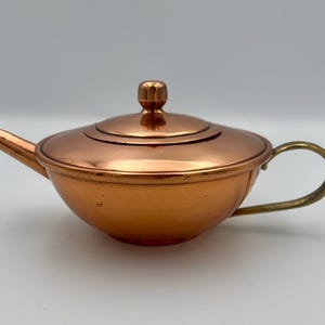 May include: A copper teapot with a lid and a brass handle. The teapot has a rounded body and a spout. The lid has a small knob on top. The handle is curved and attached to the side of the teapot. The teapot is made of copper.