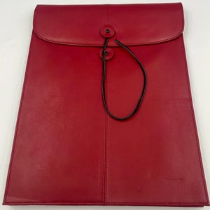 May include: A red leather portfolio with a flap closure and a black string tie. The portfolio has a vertical seam down the center and two black buttons on the flap. The overall design is sleek and professional.