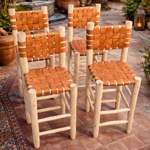 Könnte beinhalten: Fünf Holzstühle mit geflochtenen Ledersitzen und -lehnen. Die Stühle haben hellfarbene Holzrahmen und stehen auf einer Ziegelterrasse. Die Stühle sind für den Außenbereich konzipiert.