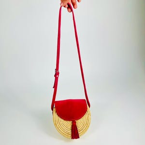 May include: A round, woven straw crossbody bag with a red suede flap and tassel. The bag has a long, adjustable red strap. The bag is held up by a hand against a white background.