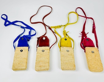 May include: Four woven straw crossbody phone bags with colorful leather accents. Each bag has a different color: blue, red, yellow, and burgundy. The bags have a rectangular shape and a long strap for wearing across the body.
