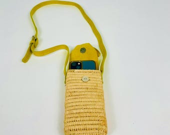 May include: A woven straw crossbody phone bag with a yellow strap and flap. The bag is rectangular with a button closure and contains a blue phone. The strap is adjustable.