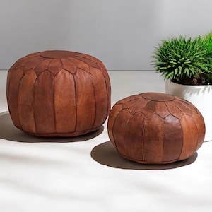 May include: Two brown leather poufs of different sizes. The larger pouf is round and has a segmented design. The smaller pouf is also round and has a similar segmented design. A potted plant is in the background.