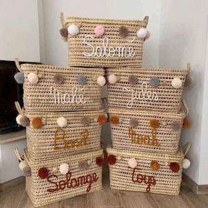 May include: A collection of woven rectangular storage baskets with handles. Each basket is adorned with pom-poms and a name or word in cursive lettering: Salome, Mahe, Jules, Basil, Solange, and toys. The baskets are beige.