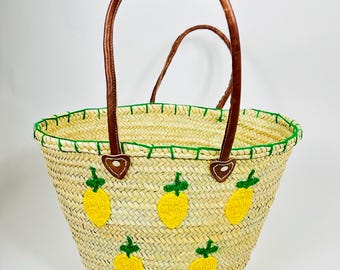 May include: A woven straw tote bag with brown leather handles and accents. The bag features embroidered yellow pineapple designs and a green stitched trim around the top edge. The bag is designed for carrying items.