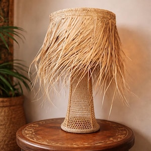 May include: A natural woven lamp with a fringed lampshade. The lamp has a cylindrical base and a flared shade, both made of woven material. The lamp sits on a round wooden table with carved details. The color palette is neutral, with beige and brown tones.
