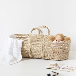 May include: A woven wicker baby bassinet with two handles, a white blanket, and a doll with blonde hair. The bassinet is light brown and has a rectangular shape. A small panda toy and a book are in the foreground.
