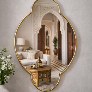 Könnte beinhalten: Goldgerahmter Wandspiegel mit einem verzierten, vierblättrigen Rahmen. Der Spiegel reflektiert ein Wohnzimmer mit Sofa, Couchtisch und architektonischen Details. Er hängt an einer hellgrauen Wand, mit einem marmorgekrönten Tisch im Vordergrund.