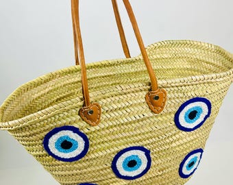 May include: Woven straw tote bag with brown leather handles and decorative blue and white evil eye designs. The bag has a natural straw colour and is held by a person.
