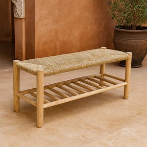 Puede incluir: Banco de madera clara con asiento tejido y estante inferior de listones. El banco tiene cuatro patas cilíndricas. El asiento tejido es de color beige claro. El banco mide aproximadamente 122 cm de largo.