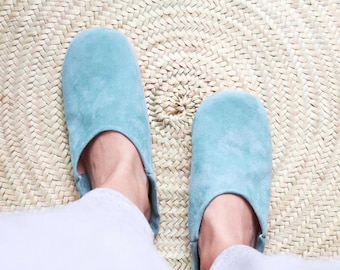 May include: A pair of light blue suede slippers. The slippers are on a woven straw mat. The slippers have a rounded toe and a low profile. The person is wearing white pants.