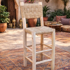 May include: A light-coloured wooden bar stool with a woven seat and backrest. The stool has a natural, rustic appearance and is placed on a patterned rug. The design is suitable for outdoor or indoor use.