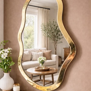 May include: A large, irregularly shaped mirror with a gold-colored frame. The mirror reflects a living room scene with a sofa, coffee table, and decorative items. The mirror is mounted on a pink wall above a green and gold console table.