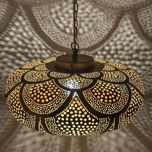 Pode incluir: Luminária pendente decorativa com acabamento em metal escuro e um intrincado design perfurado. A luz projeta uma sombra padronizada na superfície circundante. A luminária é suspensa por uma corrente.