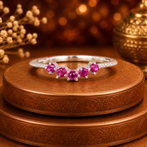May include: A silver-toned curved ring featuring five round-cut vibrant pink gemstones set in a row along the centre. The band has a delicate beaded texture along the outer edges, creating a refined and symmetrical appearance for this jewellery piece.