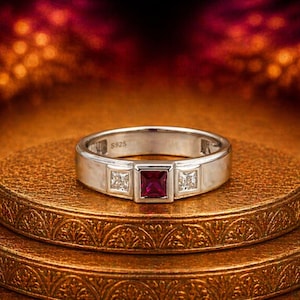 Puede incluir: Un anillo plateado con una gema roja cuadrada en el centro, flanqueada por dos piedras cuadradas claras más pequeñas. El anillo está sobre una superficie dorada texturizada. La banda está marcada con "S925".