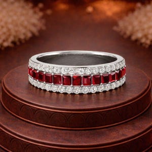 Puede incluir: Un anillo plateado con una banda de piedras rojas cuadradas, bordeada por dos filas de pequeñas piedras redondas y transparentes. El anillo se exhibe sobre una superficie marrón escalonada.