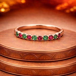May include: A rose gold ring featuring alternating green and red gemstones. The ring is set on a brown, textured surface, with a blurred, warm-toned background. The gemstones are evenly spaced around the band, creating a symmetrical design. The ring is a piece of jewelry.