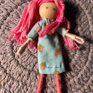 May include: A handmade doll with a wooden head and pink yarn hair. The doll wears a light blue dress with a strawberry pattern and pink yarn legs and shoes. The doll is resting on a gray crocheted surface.