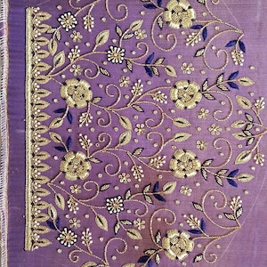 Puede incluir: Primer plano de una tela lavanda con intrincados bordados. El diseño presenta flores, hojas y vides doradas, acentuadas con hojas azul oscuro y pequeños detalles blancos. El borde tiene un borde dorado.