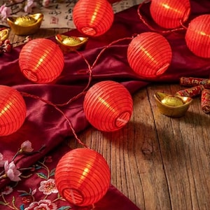 Può includere: Una stringa di lanterne di carta rosse illuminate, disposte su un tessuto rosso scuro con accenti dorati e decorazioni floreali. Le lanterne rotonde hanno un design a coste, creando un'atmosfera festosa e calda.