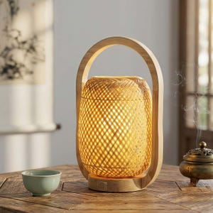 May include: A woven bamboo lantern with a wooden handle and base. The lantern emits a warm, inviting glow. A small, light green ceramic teacup and a bronze incense burner are on the wooden table.