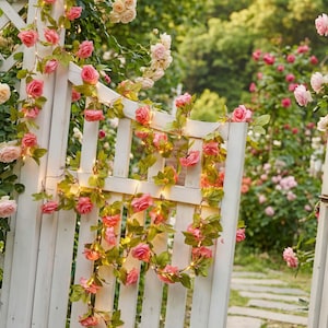 May include: A white wooden garden gate adorned with pink rose garlands and small lights. The roses are in full bloom, and the green leaves provide a natural contrast. The background features a lush garden with more roses.
