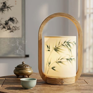 May include: A wooden lantern-style lamp with a light-colored frame and a cylindrical shade. The shade features a green bamboo leaf design. An incense burner and a small teal cup are on the wooden table. The lamp has a button on the base.