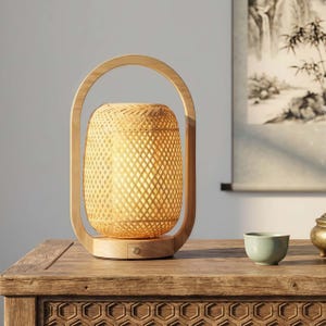 May include: A bamboo lantern with a woven shade and a wooden handle. The lamp sits on a wooden table with a small green teacup and a decorative metal container. The lamp's shade is illuminated, casting a warm glow.