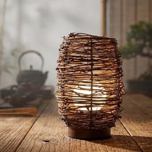 May include: A decorative table lamp with a woven, brown, twig-like shade. The lamp has a cylindrical shape and sits on a dark brown base. The light inside the shade is glowing, creating a warm ambiance. The lamp is on a wooden surface.