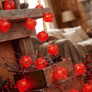 Può includere: Una stringa di luci rosse a forma di lanterna, illuminate, drappeggiate su una struttura in legno rustico. Le luci sono infilate su un filo rosso, con un bagliore caldo che emana da ogni lanterna. La scena è decorata con pigne e bacche.