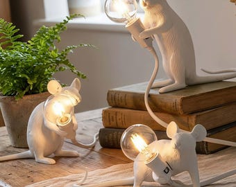May include: Three white mouse-shaped lamps with exposed light bulbs. The lamps are on a wooden table with books and a potted plant in the background. The lamps are in different poses, each holding a light bulb.